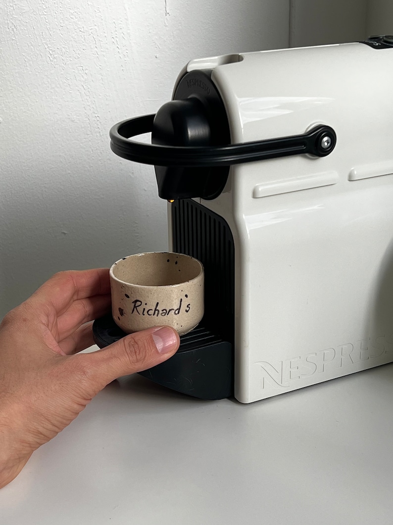 Op de afbeelding: Een witte Nespresso-koffiemachine met zwarte details. Een kleine, gespikkelde keramische beker met de tekst "Richard's" staat op de lekbak van de machine. De machine staat op een wit oppervlak.