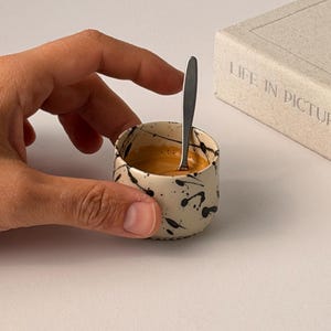Op de afbeelding: Een kleine keramische espressokop met een zwart verfspatterontwerp, gevuld met koffie en een kleine zilveren lepel. Op de achtergrond een boek met de tekst "LIFE IN PICTURE".