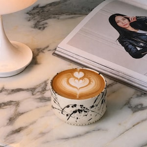 May include: A latte with heart-shaped foam art in a ceramic cup with black paint splatter design. The cup sits on a marble table next to a magazine and a white lamp.