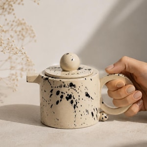 May include: A cream ceramic teapot with black splatter paint. It features a round handle, a spout, and a lid with a spherical knob. The teapot is held by a hand. The teapot is likely around 10-15 cm in height.