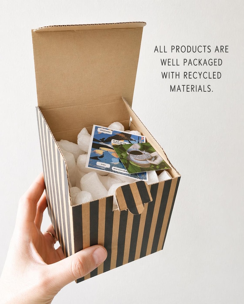 May include: A striped cardboard box with black and tan vertical stripes, open to reveal white packing peanuts and small cards. The box is held by a hand. Text on the box reads: ALL PRODUCTS ARE WELL PACKAGED WITH RECYCLED MATERIALS.