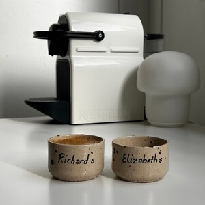 Op de afbeelding: Twee kleine, gespikkelde keramische koffiekopjes, waarvan &eacute;&eacute;n gevuld met koffie, staan voor een witte Nespresso-machine. Op de linkerbeker staat "Richard's" en op de rechter "Elizabeth's". Rechts staat een witte paddenstoelvormige lamp.