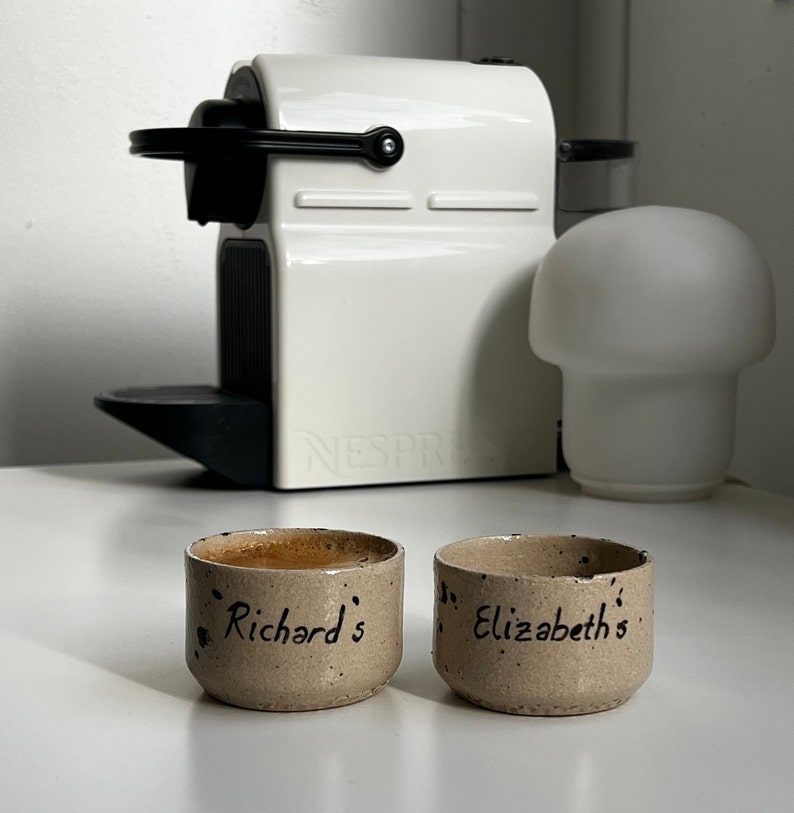 Op de afbeelding: Twee kleine, gespikkelde keramische koffiekopjes, waarvan &eacute;&eacute;n gevuld met koffie, staan voor een witte Nespresso-machine. Op de linkerbeker staat "Richard's" en op de rechter "Elizabeth's". Rechts staat een witte paddenstoelvormige lamp.