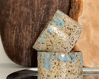 May include: Two ceramic cups with a speckled brown glaze and blue accents. The cups are stacked on top of each other and are sitting on a silver tray.