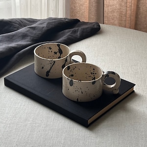 May include: Two handmade ceramic mugs with a speckled, off-white finish and black paint splatters. Each mug has a rounded handle with a small, decorative ball. The mugs are placed on a black book.