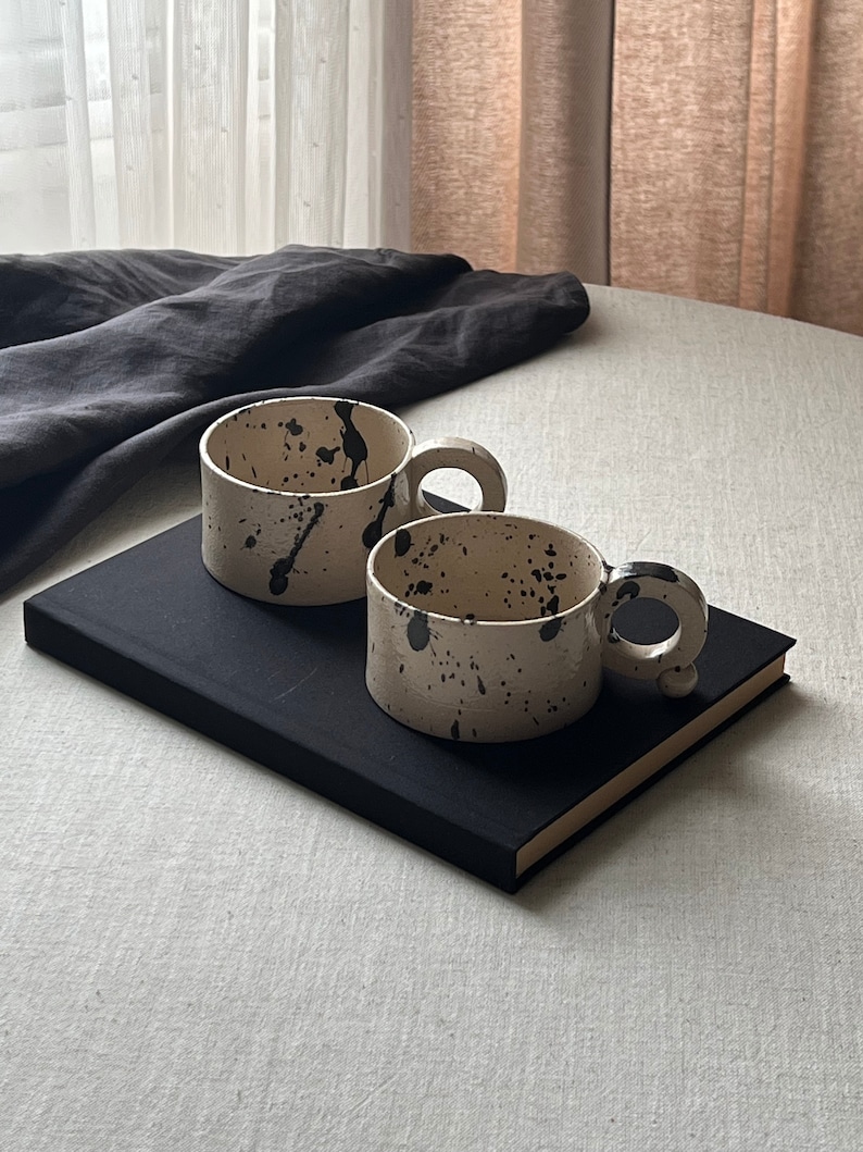 May include: Two handmade ceramic mugs with a speckled, off-white finish and black paint splatters. Each mug has a rounded handle with a small, decorative ball. The mugs are placed on a black book.