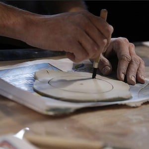 Op de afbeelding: Close-up van handen die een gereedschap gebruiken om een cirkelvormig ontwerp in een stuk klei te snijden. De klei is lichtbruin en ligt op een bedrukt oppervlak. Een ambachtelijk project in uitvoering.
