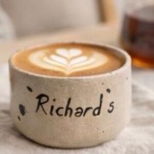 Op de afbeelding: Een beige keramische koffiekop met de naam "Richard's" op de zijkant. De kop bevat een latte met een hartvormig ontwerp in het schuim. De kop staat op een witte doek.