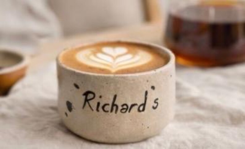 Op de afbeelding: Een beige keramische koffiekop met de naam "Richard's" op de zijkant. De kop bevat een latte met een hartvormig ontwerp in het schuim. De kop staat op een witte doek.