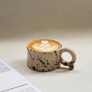 May include: A ceramic coffee mug with a speckled design, filled with a latte featuring a heart-shaped foam design. The mug has a unique handle and sits next to an open magazine.
