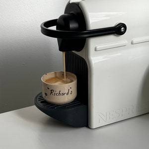 Op de afbeelding: Een witte Nespresso-koffiemachine met een zwarte handgreep en tuit die koffie uitschenkt in een kleine, gespikkelde keramische beker. De beker is voorzien van de naam "Richard's". De machine staat op een wit oppervlak.