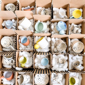 May include: Assortment of ceramic mugs and bowls in cardboard boxes, packed with white foam. The mugs and bowls are various colors, including white, yellow, blue, and speckled designs. Some have handles, and others are handleless.