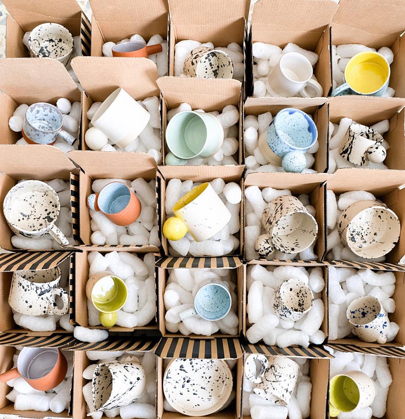 May include: Assortment of ceramic mugs and bowls in cardboard boxes, packed with white foam. The mugs and bowls are various colors, including white, yellow, blue, and speckled designs. Some have handles, and others are handleless.