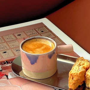 Pode incluir: Uma caneca de cerâmica rosa e azul cheia de café, colocada numa bandeja prateada com biscoitos. A caneca tem uma pega e uma base clara. O café tem uma camada de espuma. A bandeja está sobre uma superfície com um tablet e uma revista.