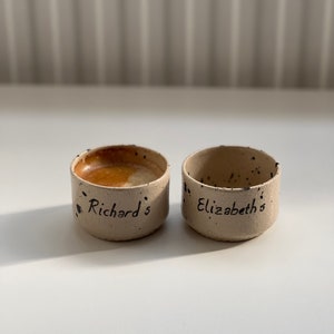 Op de afbeelding: Twee kleine, handgemaakte keramische kommen met een gespikkelde, beige buitenkant. E&eacute;n kom is gevuld met een bruine vloeistof. De kommen zijn beschreven met de namen "Richard's" en "Elizabeth's" in zwart schrift.