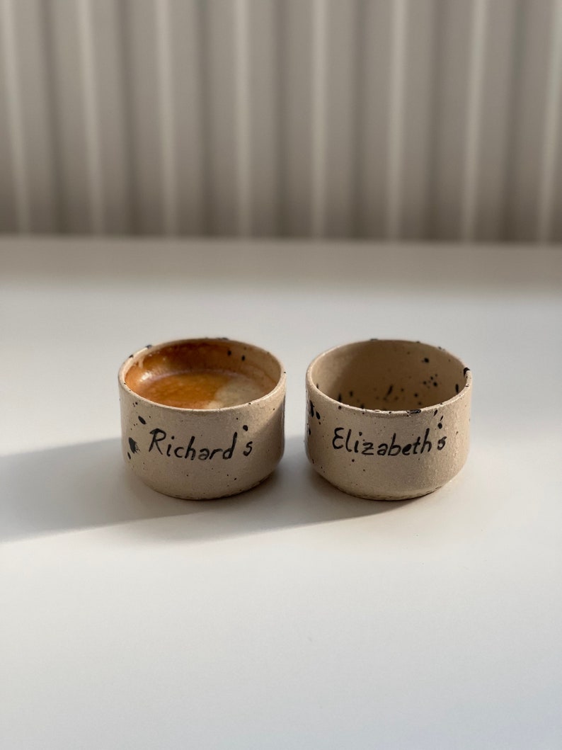Op de afbeelding: Twee kleine, handgemaakte keramische kommen met een gespikkelde, beige buitenkant. E&eacute;n kom is gevuld met een bruine vloeistof. De kommen zijn beschreven met de namen "Richard's" en "Elizabeth's" in zwart schrift.