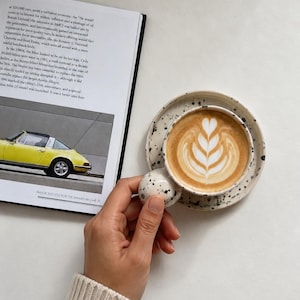 May include: A latte in a speckled ceramic cup with a distinctive handle, on a matching saucer. The coffee has latte art. An open book with a yellow car image is visible.