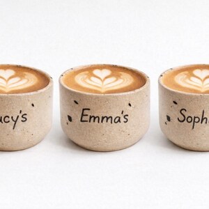 Op de afbeelding: Drie beige keramische koffiekopjes met latte art. Elke beker heeft een naam: "Lucy's", "Emma's" en "Sophie's". De koffie heeft een hartvormig ontwerp.