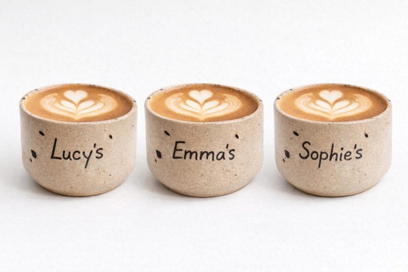 Op de afbeelding: Drie beige keramische koffiekopjes met latte art. Elke beker heeft een naam: "Lucy's", "Emma's" en "Sophie's". De koffie heeft een hartvormig ontwerp.