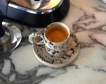 May include: A ceramic espresso cup and saucer set with a speckled black and white design. The cup is filled with a dark brown espresso. The set is on a marble surface, with a coffee machine and tamper visible in the background.