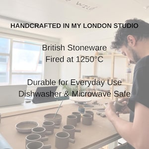 May include: A collection of handcrafted, gray stoneware mugs with handles, arranged on a wooden table. The text "HANDCRAFTED IN MY LONDON STUDIO" is visible. The mugs are durable for everyday use and are dishwasher and microwave safe.