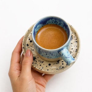 May include: A ceramic espresso cup and saucer set. The cup is blue with a speckled design and a handle. The saucer is beige with black speckles. The cup is filled with espresso. The set is held in a hand.