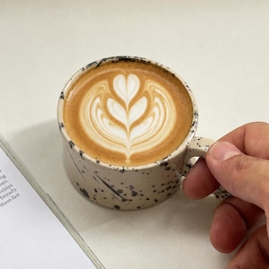 May include: A ceramic mug with a speckled beige exterior and a handle, filled with a latte featuring a heart-shaped design in the foam. A book with text is visible in the background.