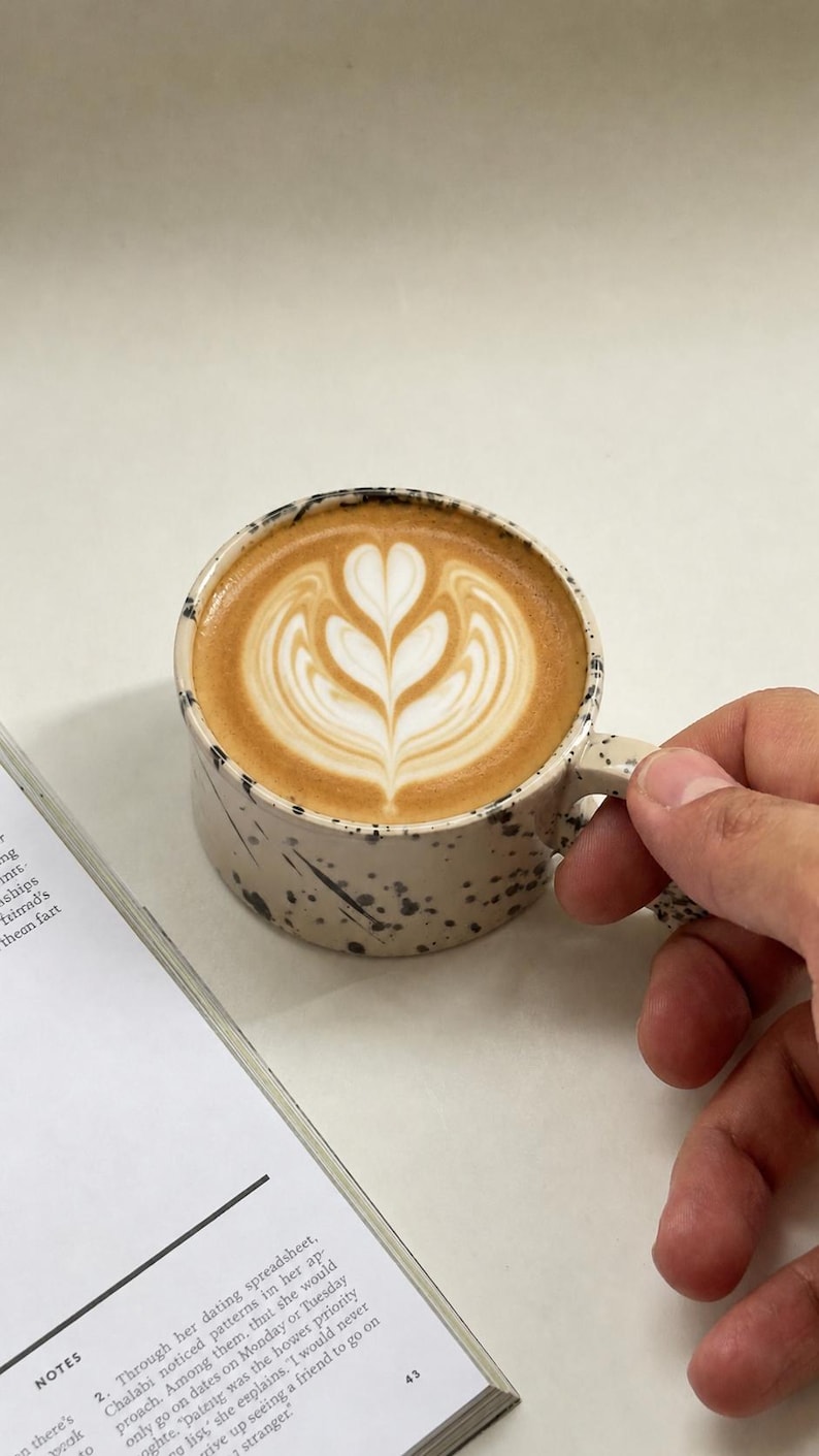 May include: A ceramic mug with a speckled beige exterior and a handle, filled with a latte featuring a heart-shaped design in the foam. A book with text is visible in the background.