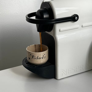 Op de afbeelding: Een witte Nespresso koffiemachine die koffie uitschenkt in een kleine, gespikkelde keramische beker. De beker is voorzien van de naam "Richard's". De machine heeft een zwarte handgreep en accenten, en de merknaam is zichtbaar.