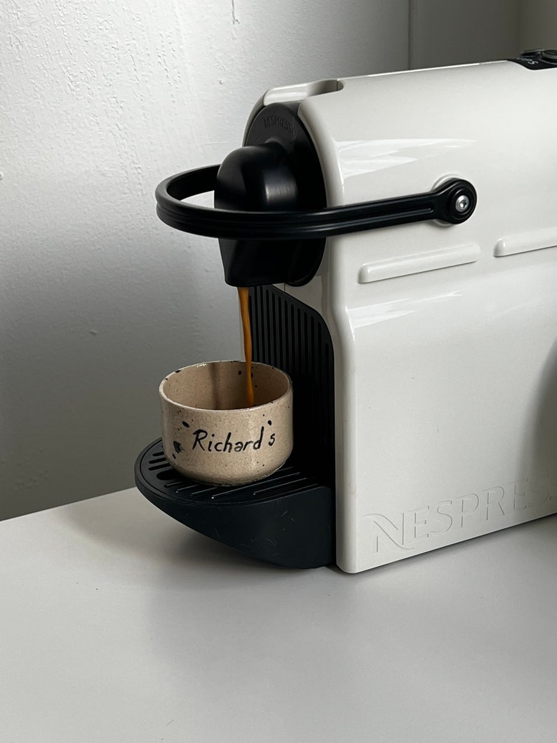 Op de afbeelding: Een witte Nespresso koffiemachine die koffie uitschenkt in een kleine, gespikkelde keramische beker. De beker is voorzien van de naam "Richard's". De machine heeft een zwarte handgreep en accenten, en de merknaam is zichtbaar.