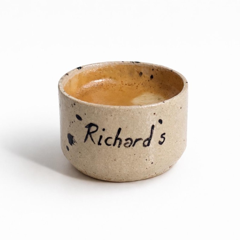 Op de afbeelding: Een kleine, beige keramische beker met de naam "Richard's" in zwart schrift. De beker is gevuld met een lichtbruine vloeistof, waarschijnlijk koffie of espresso. De beker heeft donkerblauwe spikkels.