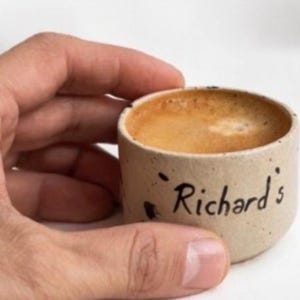 Op de afbeelding: Een kleine, beige keramische espressokop gevuld met koffie, vastgehouden door een hand. Op de kop staat in zwarte letters "Richard's". De koffie heeft een lichtbruine crema.