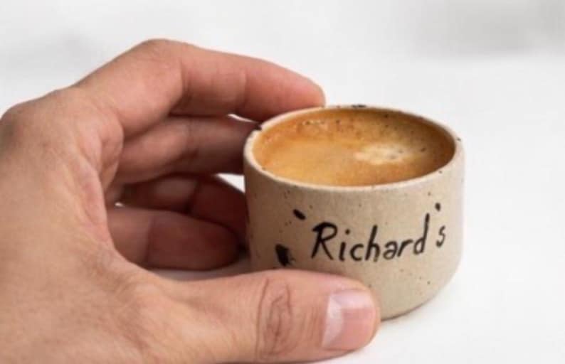 Op de afbeelding: Een kleine, beige keramische espressokop gevuld met koffie, vastgehouden door een hand. Op de kop staat in zwarte letters "Richard's". De koffie heeft een lichtbruine crema.