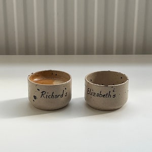 Op de afbeelding: Twee kleine, handgemaakte keramische bekers met een gespikkelde, beige afwerking. E&eacute;n beker bevat een donkere vloeistof. De bekers zijn beschreven met de namen "Richard's" en "Elizabeth's" in zwart schrift.