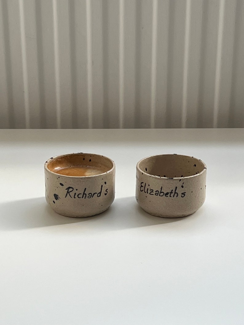 Op de afbeelding: Twee kleine, handgemaakte keramische bekers met een gespikkelde, beige afwerking. E&eacute;n beker bevat een donkere vloeistof. De bekers zijn beschreven met de namen "Richard's" en "Elizabeth's" in zwart schrift.