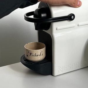 Op de afbeelding: Een witte Nespresso-koffiemachine met zwarte details. Een kleine, gespikkelde keramische beker met de tekst "Richard's" staat op de lekbak. De machine staat op een wit oppervlak.