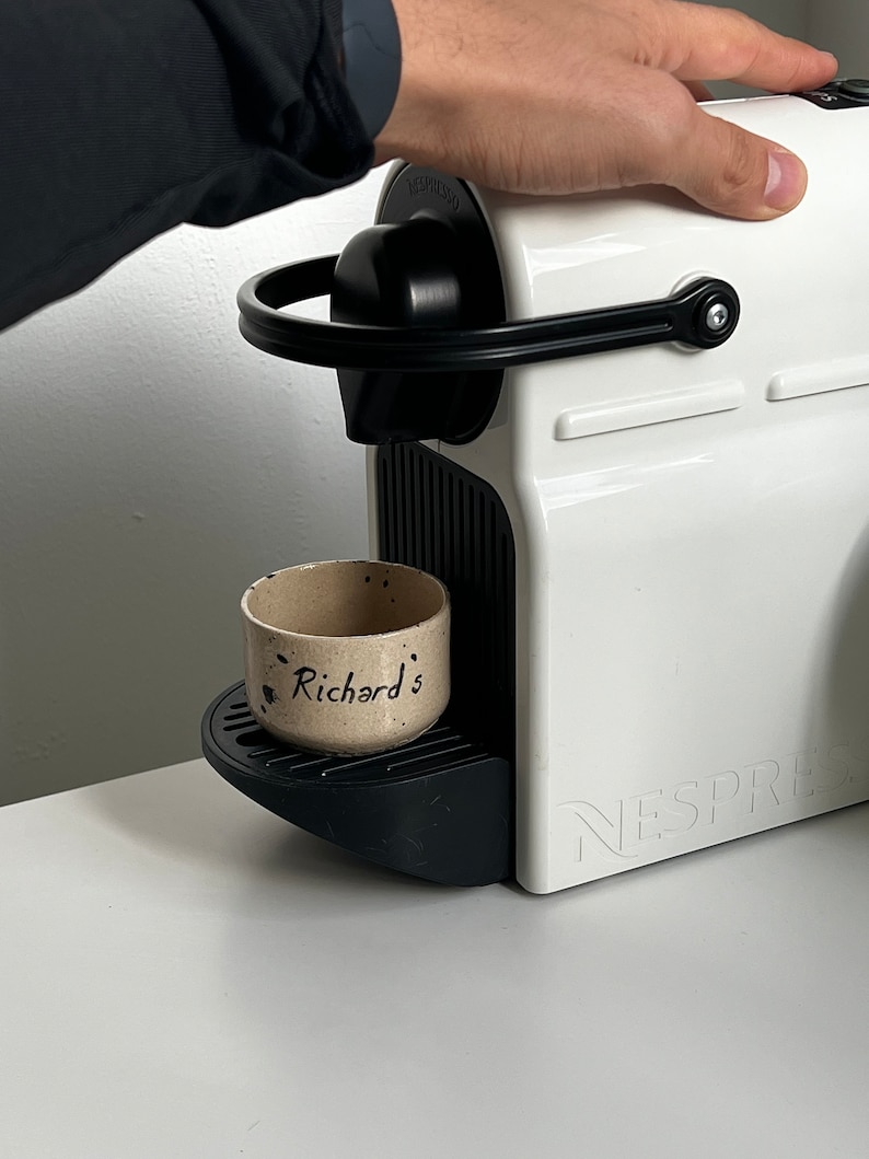 Op de afbeelding: Een witte Nespresso-koffiemachine met zwarte details. Een kleine, gespikkelde keramische beker met de tekst "Richard's" staat op de lekbak. De machine staat op een wit oppervlak.