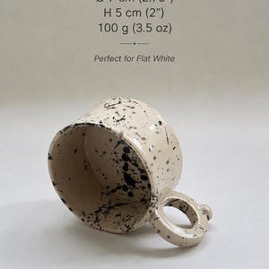 May include: A beige ceramic mug with black splatter details. The mug has a rounded handle and is 2 inches high, 2.75 inches in diameter, and weighs 3.5 oz. The text on the image reads "5 oz / 150 ml" and "Perfect for Flat White."