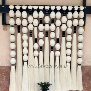 May include: A wall hanging made of white yarn with black accents. The yarn is strung together in a series of loops and knots, creating a unique and modern design. The wall hanging is hung on a black wooden frame. The text "SN HANDICRAFTS" is visible in the image.