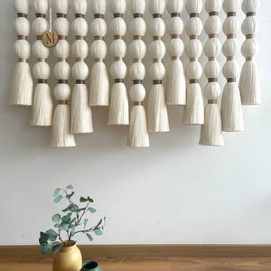 May include: A large, cream-colored wall hanging with a repeating pattern of round, beaded elements and tassels. The hanging is suspended from a chain. A small wooden disc with the letter "M" hangs in the center. A yellow vase and a green vase are on the floor.