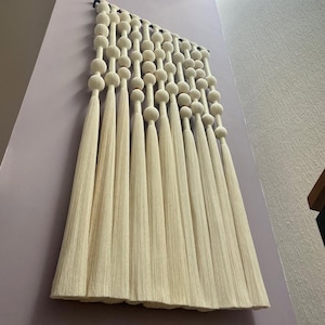 May include: Cream-coloured macrame wall hanging featuring a series of large, round beads and long, flowing tassels. The wall decor is mounted on a light purple wall. The design is a modern, bohemian style.