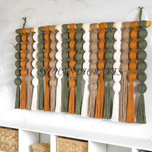 May include: A wall hanging made of yarn with a wooden dowel at the top. The yarn is woven into a series of balls and tassels in shades of green, brown, and white. The wall hanging is hanging on a white wall above a white shelf with woven baskets.