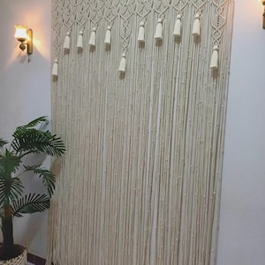 May include: Cream-colored macrame curtain with a geometric top design and hanging tassels. The curtain is made of thick, woven cotton cords, creating a textured, bohemian look. The curtain is hung on a wooden rod.