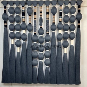 Könnte beinhalten: Ein Wandbehang aus grauem Garn mit schwarzen und goldenen Akzenten. Das Garn ist zu einer Reihe von Knoten und Schlaufen gebunden, was ein strukturiertes und optisch ansprechendes Design ergibt. Der Wandbehang ist etwa 76 cm lang und 38 cm breit.