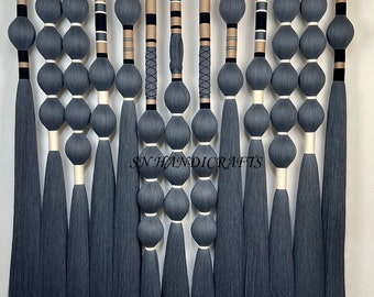 May include: A handcrafted wall hanging featuring multiple strands of gray yarn. Each strand has a series of gray, spherical shapes and a long tassel. Some strands have decorative elements in black, beige, and white. The text "SN HANDICRAFTS" is visible.