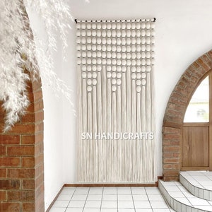 Puede incluir: Un tapiz de macramé blanco con un patrón geométrico de pompones blancos y largos hilos de lana. El tapiz está montado en una pared blanca con un arco de ladrillo en el fondo. El texto "SN HANDICRAFTS" es visible en el centro inferior de la imagen.