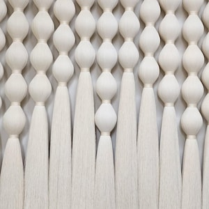 Könnte beinhalten: Cremefarbener Wandbehang mit einem sich wiederholenden Muster aus abgerundeten Formen und langen, fließenden Quasten. Das Design zeigt eine Reihe von verbundenen, bauchigen Formen, die jeweils mit einem dünnen Band gebunden sind und zu den gefransten Enden führen.