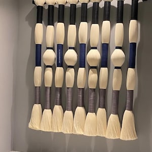 May include: A decorative wall hanging featuring eleven vertical brush-like elements. Each element has a cream-colored brush head, a black or gray handle, and sections wrapped in navy blue or gray. The elements are mounted on a black metal bar.