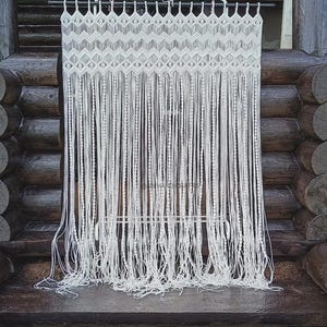 Puede incluir: Una cortina de macramé blanca con un diseño geométrico en la parte superior y largas franjas verticales. La cortina está suspendida de una barra y cuelga frente a una estructura de madera. El macramé está hecho de un material grueso y tejido.