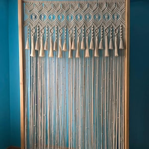 Puede incluir: Un tapiz de macramé de pared con un marco de madera y borlas. El macramé está hecho de cordón de color natural y tiene un patrón geométrico.